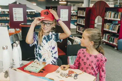 To jenter på Kongsberg bibliotek prøver caps og jojoer de har vunnet i Sommerles.